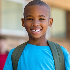 A young boy wearing a blue shirt and backpack smiles after receiving a metal-free dental crown