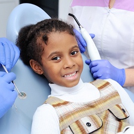 A little girl wearing a plaid jumper smiles in preparation for a regular dental checkup and cleaning