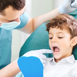 A dentist examines a young boy’s mouth while he looks in a handheld mirror