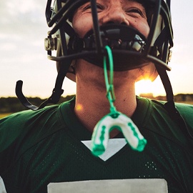 A young boy wearing football pads has a customized sportsguard hanging from his helmet