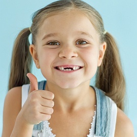 A little girl wearing denim overalls and pigtails gives a thumb up while smiling and missing her upper two front teeth
