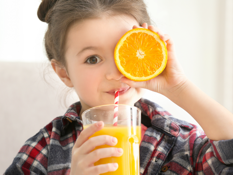 Little girl drinking orange juice and holding up orange slice over eye