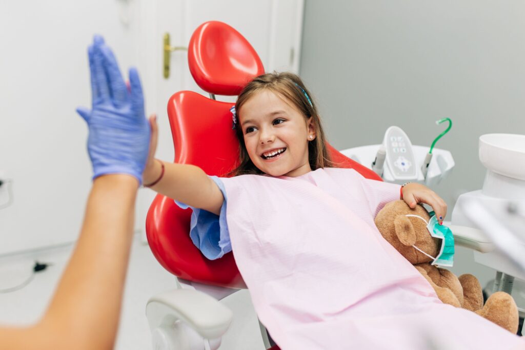 Little girl in dental chair with teddy bear high fiving dentist